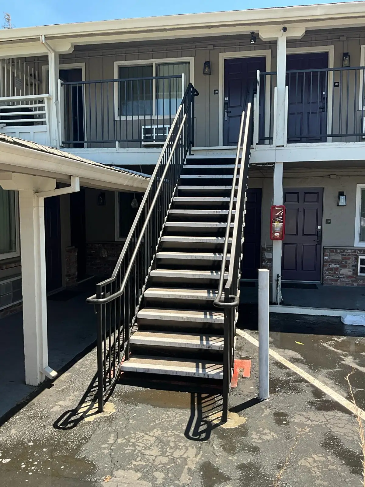 Two-story motel exterior with outdoor black metal stairs leading to upper-floor rooms and parking spaces.