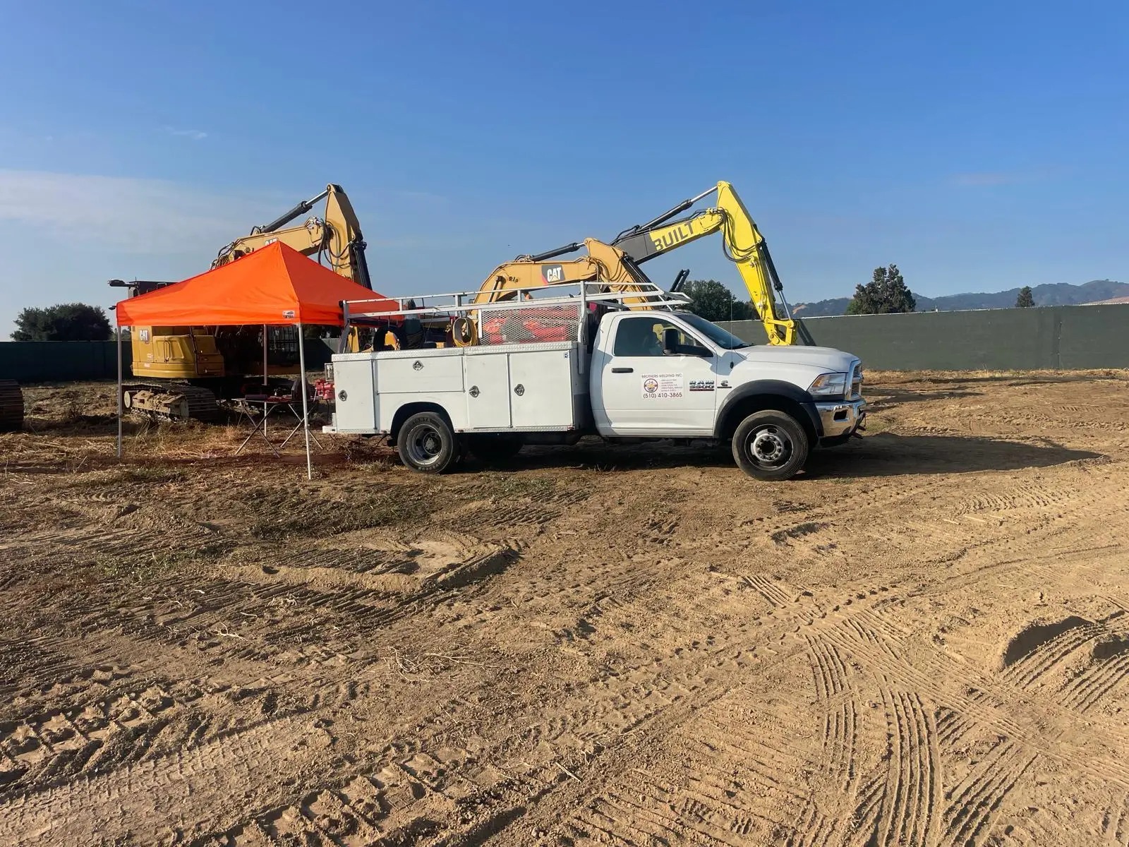 White utility truck parked beside an orange canopy tent and yellow excavators at