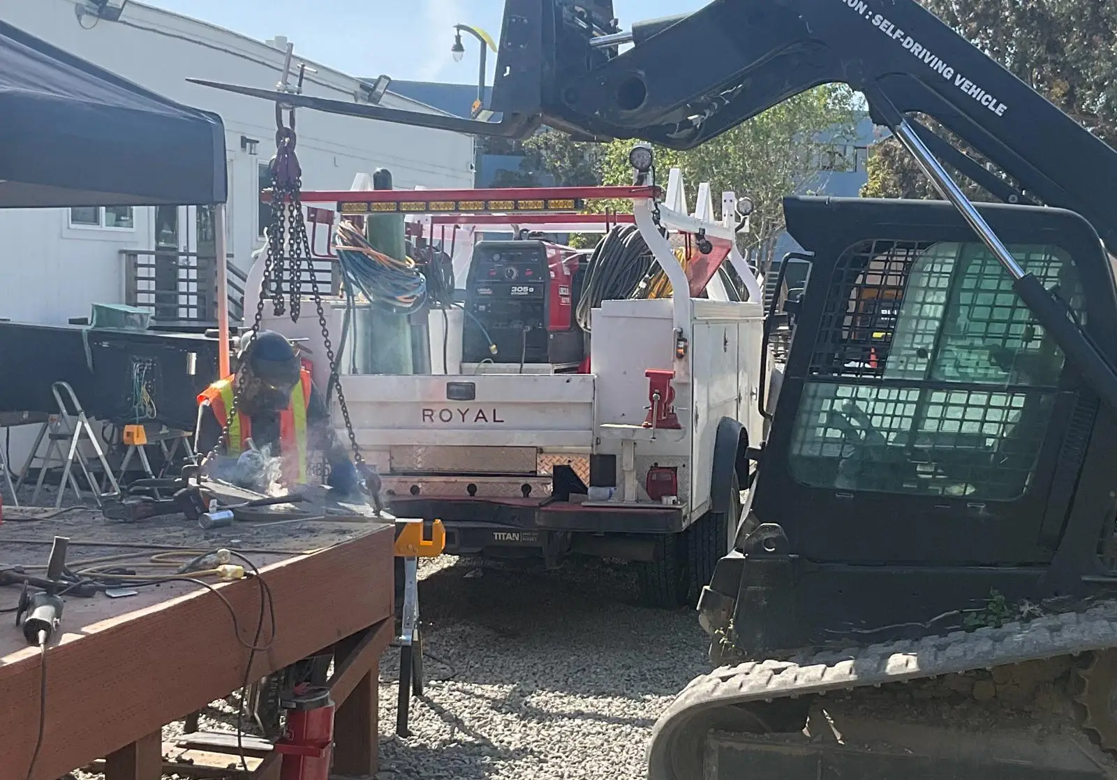Worker welding metal beside a service truck and heavy equipment at an outdoor construction site.