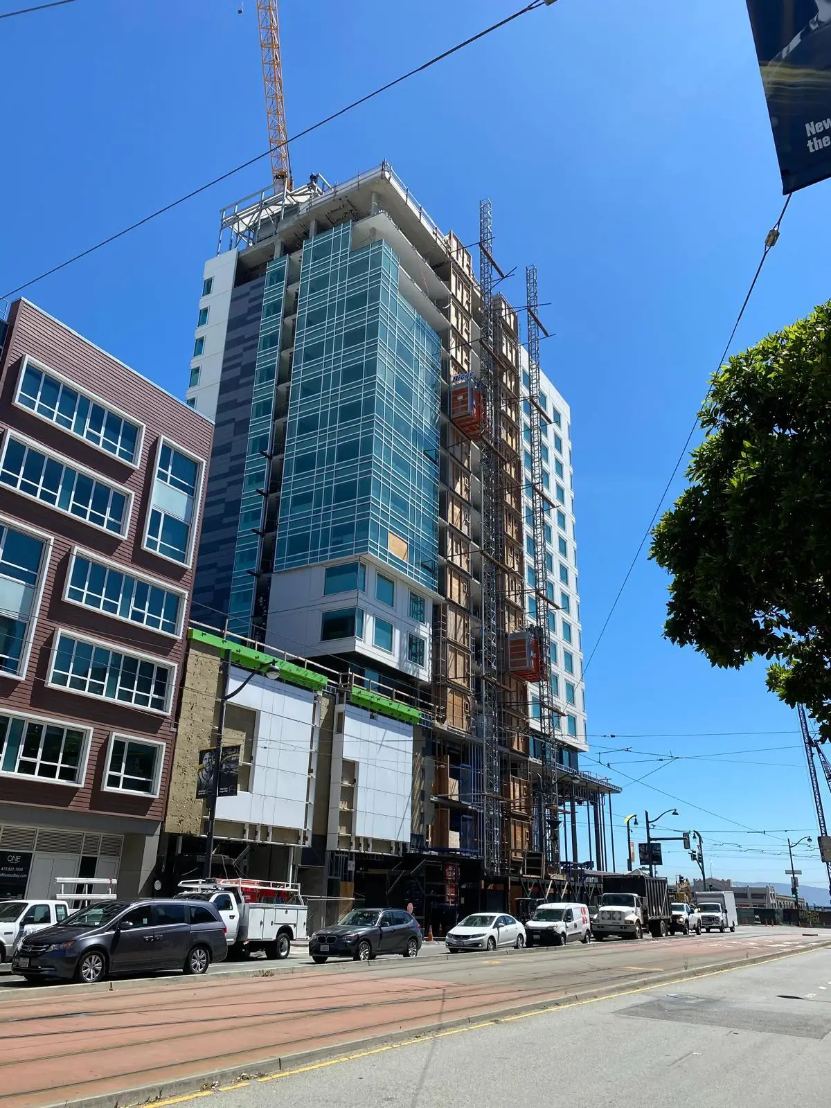 Modern high-rise building under construction with glass facade, crane, and scaffolding along a city street.