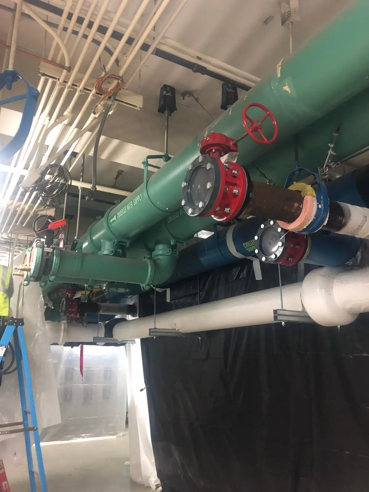 Industrial condenser water supply pipes and red control valves suspended from the ceiling of a mechanical room.