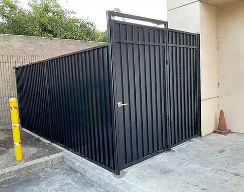 Black metal gate enclosing a storage area beside a building, with a yellow post and traffic cone nearby.