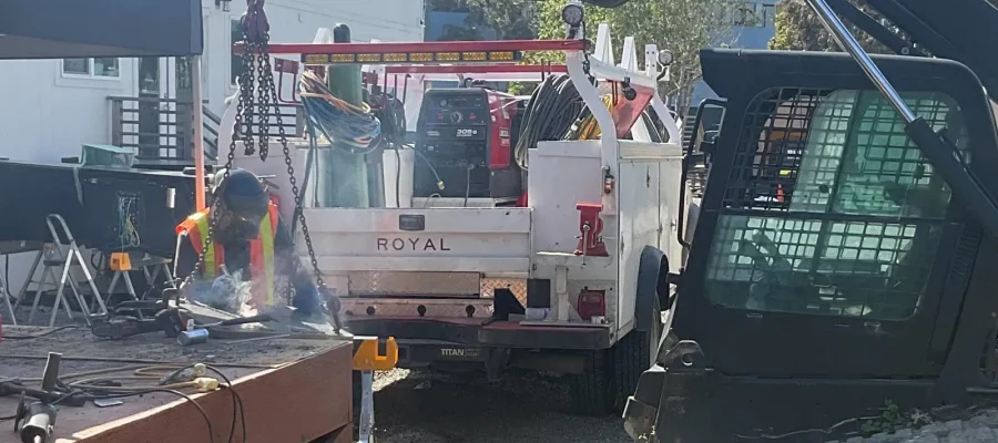 Worker welding metal beside a service truck and heavy equipment at an outdoor construction site.
